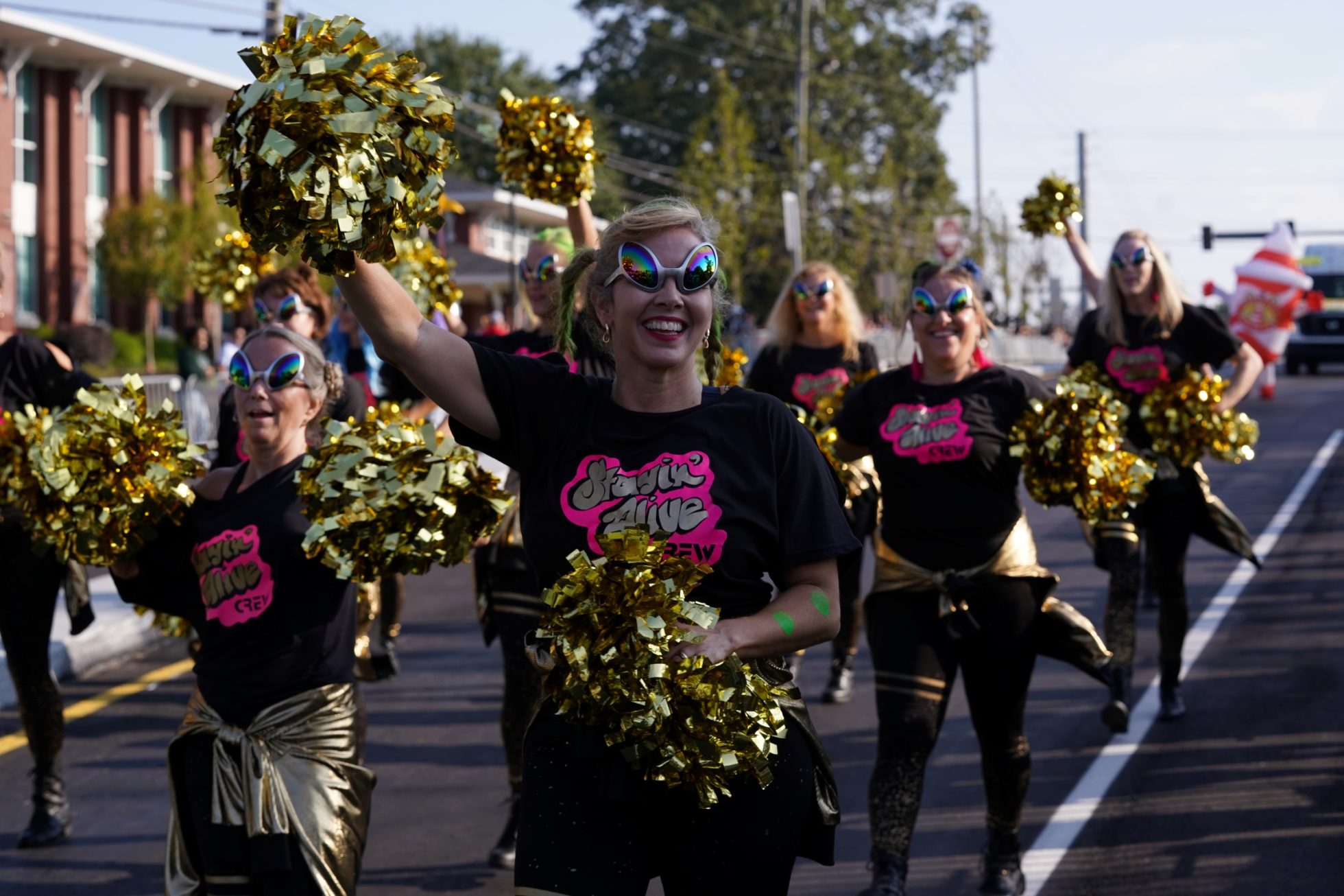 Group of people in the Suwanee Fest parade wearing black t-shirts and gold pants, holding gold pom-poms and wearing alien-like sunglasses.