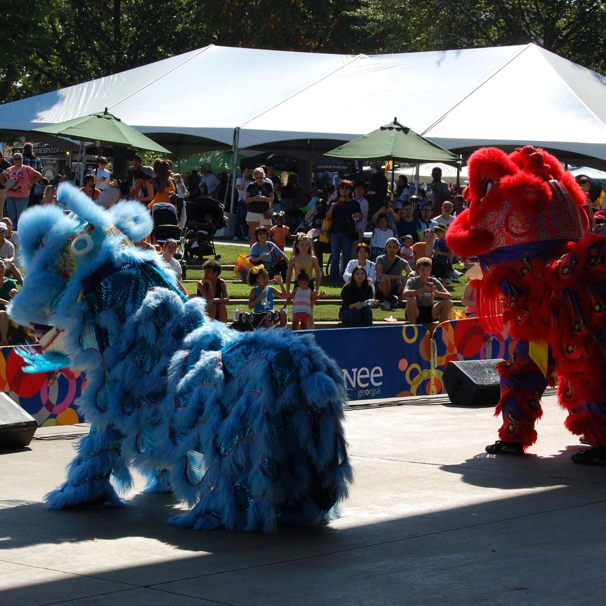 Two performers in blue and red dragon costumes dance on the Suwanee Fest stage with a crowd watching in the background.