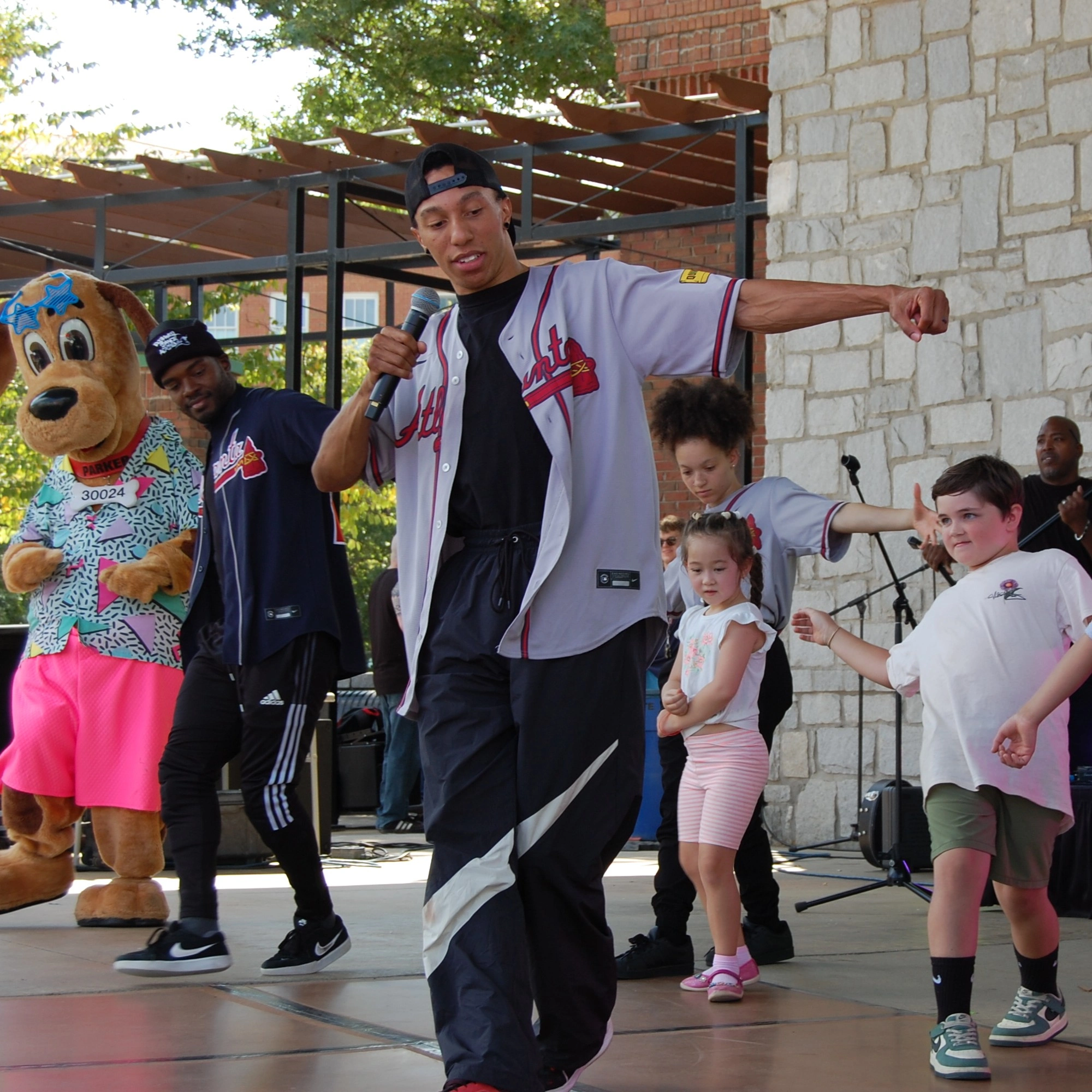 A group of people, including a man dancing with a microphone and Parker, a dog mascot, perform on an outdoor stage.