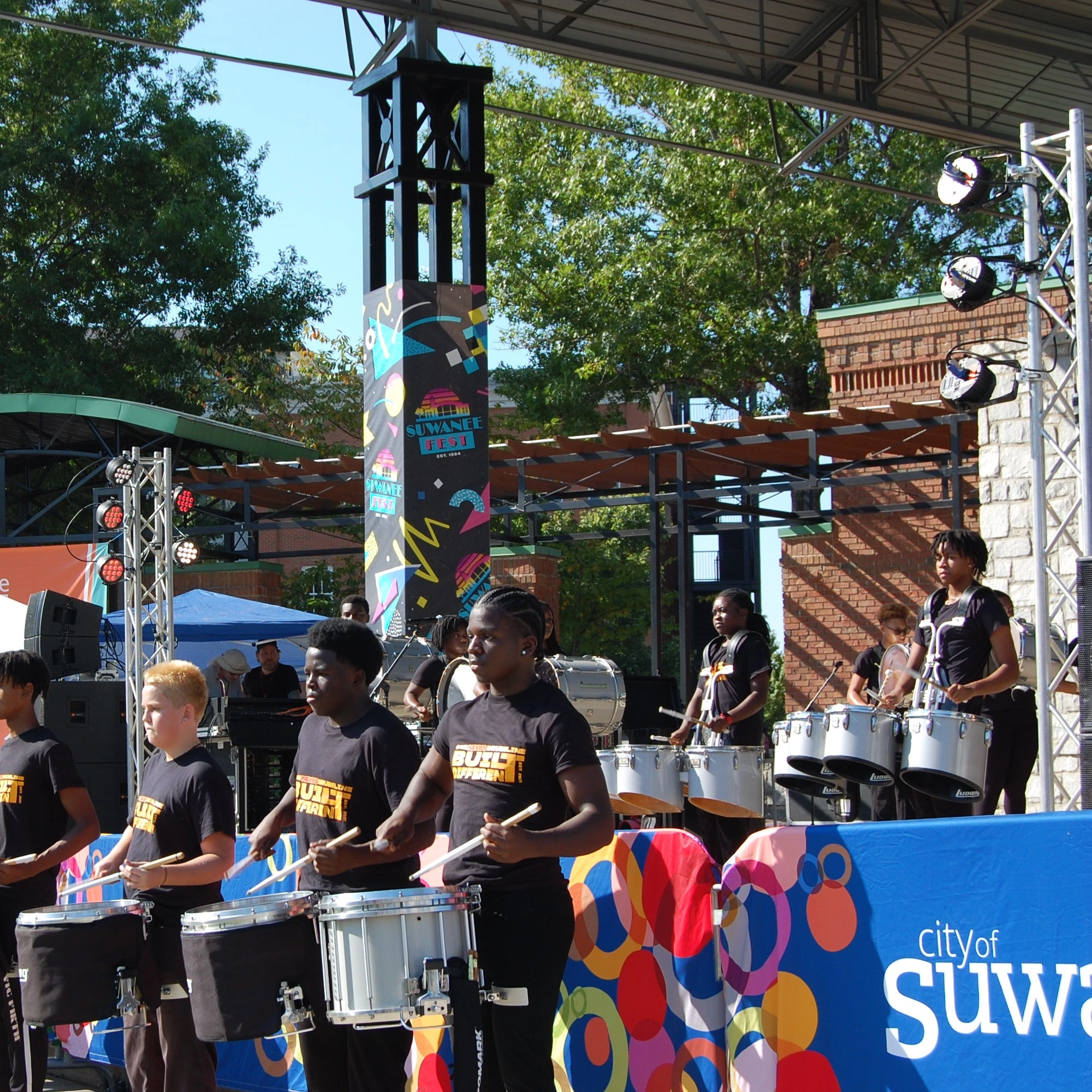 Young band playing percussion instruments on an outdoor stage with colorful decorations.