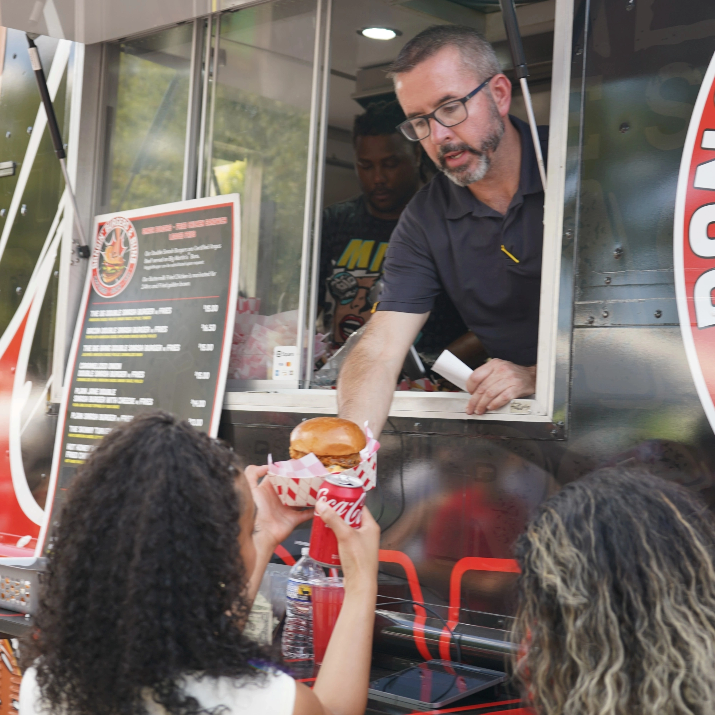 A man hands a burger to a woman at a food truck window. Another person is visible behind the counter. The customer, facing away, holds a drink in her other hand.