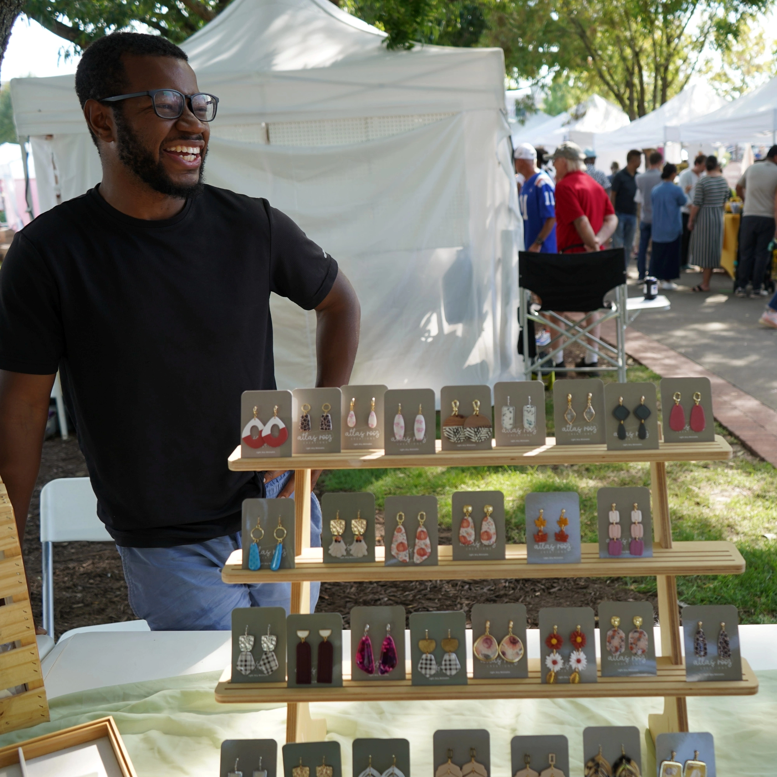 Man standing next to a display of earrings at Suwanee Fest, with tents and people in the background.