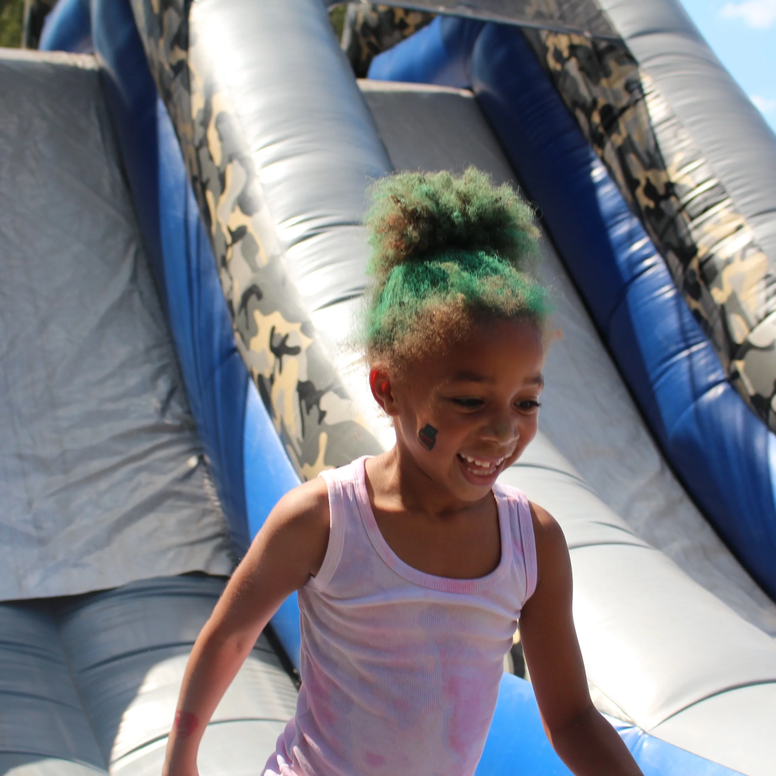 Child with green hair smiling on an inflatable slide.