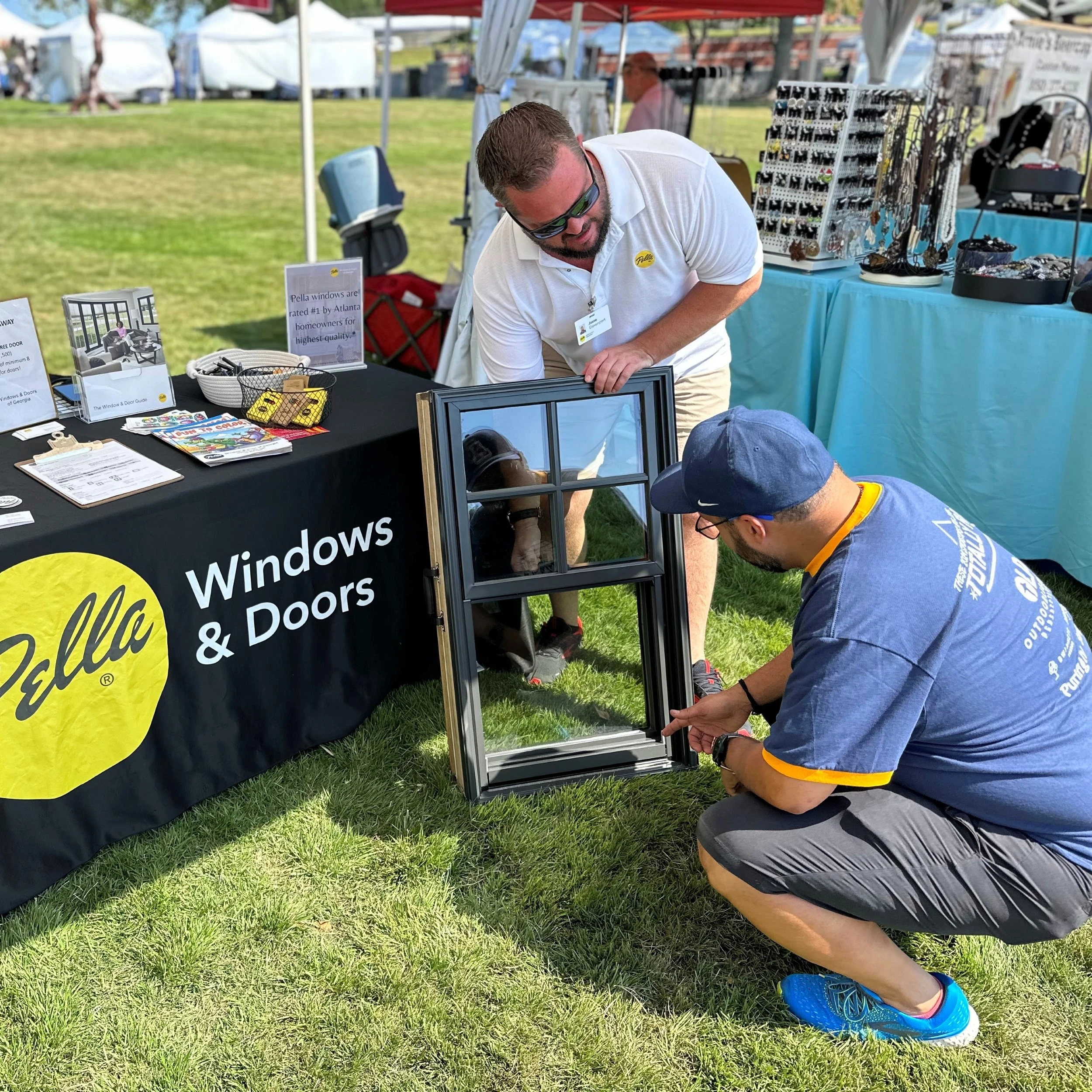 Two men examine a window sample at a sponsor booth with tables displaying brochures and swag.